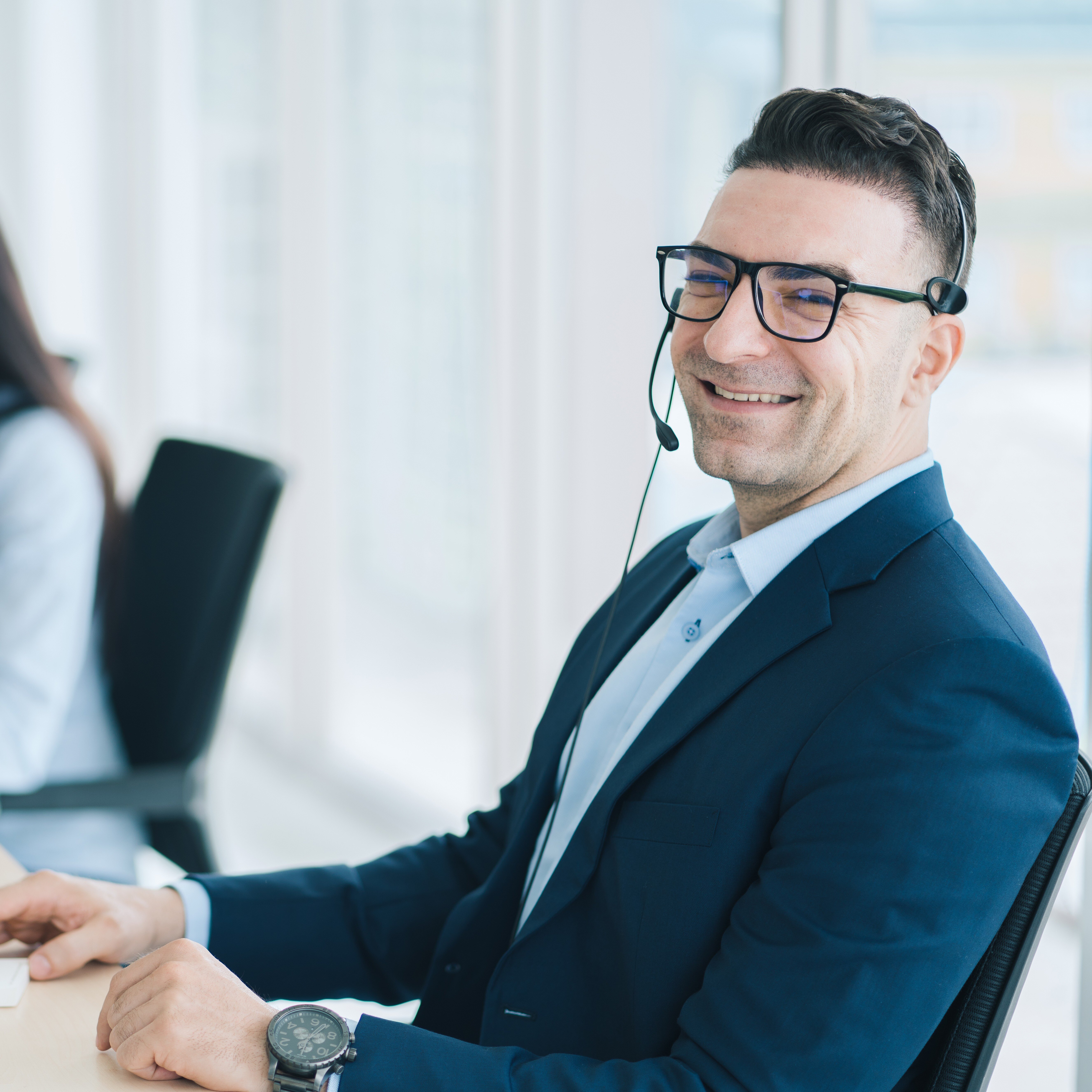 An agent in a blue blazer and black frame glasses sitting at his work station smiling. 