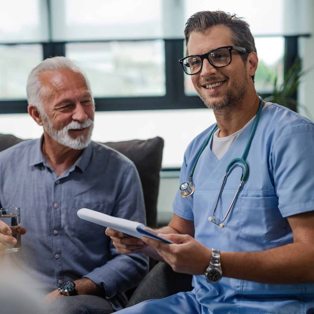 A middle-aged male clinician wearing blue scrubs, a stethoscope, and glasses is smiling while holding a patient chart. He is seated next to an older male patient with gray hair and a beard, who is also smiling and holding a glass of water. They are in a relaxed, informal setting, at hospital with natural light coming through large windows in the background.